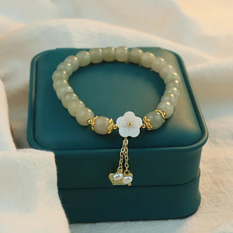 Sage-green beaded bracelet with pale flower detail and gold-tone dangling chains displayed on a green box