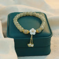Sage-green beaded bracelet with pale flower detail and gold-tone dangling chains displayed on a green box