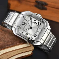 A stainless steel analog watch with a square bezel, silver-tone metal bracelet, and a white dial with black Roman numerals, resting on a vintage book.