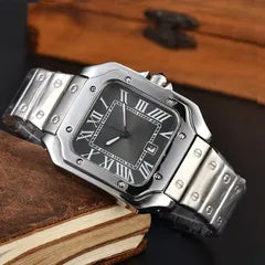 A silver-toned metal watch with a black dial and Roman numeral hour markers, resting on a stack of vintage books.