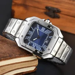 A stainless steel men's watch with a square blue dial and silver-tone metal bracelet, styled on a dark surface with a wooden box and books in the background.