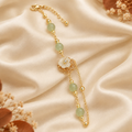 A gold-toned beaded bracelet with light green jade-like beads, a white flower centerpiece, and delicate chain extensions, styled on draped cream satin fabric with dried floral accents.
